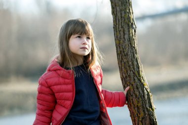 Portrait of a pretty child girl standing near a tree trunk in autumn outdoors.
