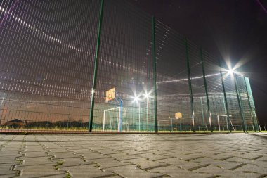 Açık havada top kapısı ve sepetli mini futbol ve basketbol sahası gece spot lambalarıyla aydınlatılmış yüksek korumalı çitlerle çevrili..