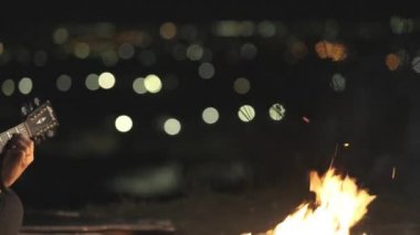 Genç, güzel bir müzisyen kadın gecenin körelmiş şehir ışıklarının arka planında şenlik ateşi yakınında gitar çalıyor..