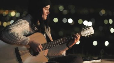 Genç, güzel bir müzisyen kadın gecenin körelmiş şehir ışıklarının arka planında şenlik ateşi yakınında gitar çalıyor..