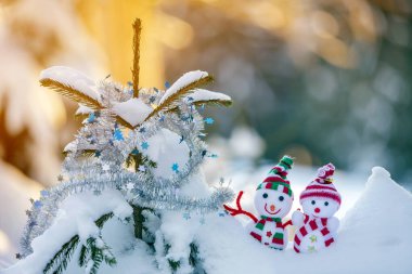 İki komik oyuncak bebek kardan adam örgü şapkalı ve eşarplı açık havada, çam ağacı dalının yanında. Mutlu yıllar ve Mutlu Noeller tebrik kartı.