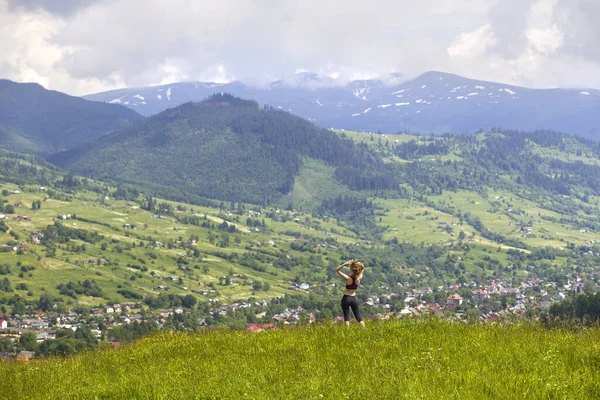 Slim young woman with raised arms outdoors on background of beautiful mountain landscape on sunny summer day.