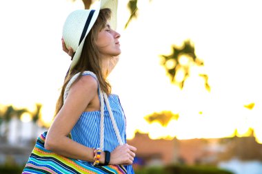 Açık mavi yaz elbisesi ve sarı hasır şapka giyen genç bir kadın şık bir omuz çantası tutuyor yaz parkında gün batımında sıcak havanın tadını çıkarıyor..