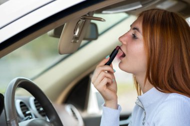 Closeup of a young redhead woman driver correcting her makeup with dark red lipstick looking in car rearview mirror behind steering wheel of a vehicle. clipart