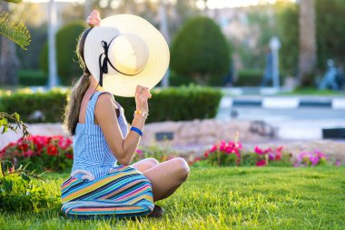 Açık mavi yaz elbisesi ve sarı hasır şapka giyen genç bir kadın yaz parkında yeşil çimlerde dinleniyor. Rahat giyimli bir kız dışarıda dinleniyor sıcak bir sabahta boş zamanın tadını çıkarıyor..