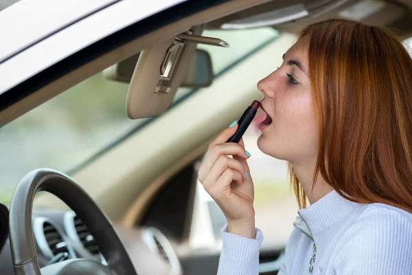 若い赤頭の女性ドライバーの閉鎖彼女のメイクを修正暗い赤口紅で車の後ろに鏡を見ますステアリングホイールの後ろ — ストック写真