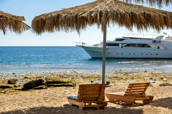 Wooden deck chairs under rough straw sun umbrella on sea beach and big white yacht ship in water near shore on sunny summer day.