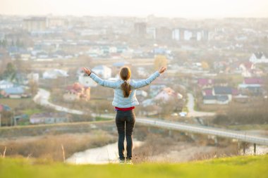 Dışarıda duran genç bir kadın şehir manzarasının tadını çıkarırken ellerini kaldırıyor. Rahatlama, özgürlük ve sağIık kavramı.