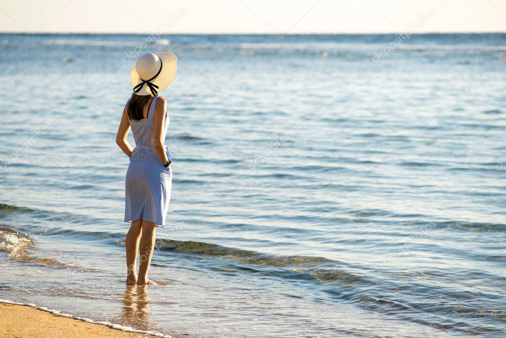 Young woman in straw hat and a dress standing alone on empty sand beach at sea shore. Lonely tourist girl looking at horizon over calm ocean surface on vacation trip.