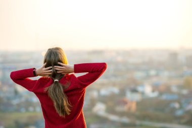 Kırmızı ceketli genç bir kadın ellerini başının arkasına koyup akşam manzarasının keyfini çıkarıyor. Rahatlama, özgürlük ve sağIık kavramı.