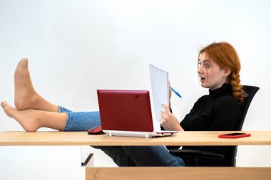 Mutlu genç ofis çalışanı ayakları çalışma masasının arkasında, dizüstü bilgisayarı, cep telefonu ve not defteriyle rahat bir şekilde oturuyor..