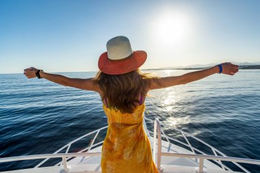 Uzun saçlı, sarı elbiseli, hasır şapkalı, beyaz yatlı güvertede duran ve mavi deniz suyunun keyfini çıkaran genç bir kadın..