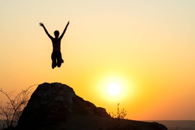 Gün batımında dağlarda boş kayalarda tek başına atlayan bir kadın yürüyüşçü silueti. Kadın turist, akşam vakti elini kaldırarak uçurumun kenarında duruyor. Turizm, seyahat ve sağlıklı yaşam tarzı konsepti.