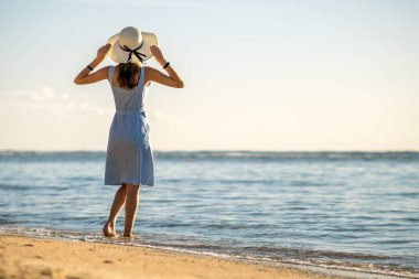 Hasır şapkalı genç bir kadın ve deniz kıyısındaki boş kumsalda tek başına duran bir elbise. Yalnız turist kız, sakin okyanus yüzeyinden ufka bakıyor. Tatilde..