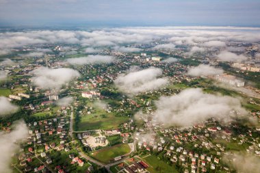 Yazın yeşil tarlalar arasında sıralı binalar ve kıvrımlı sokaklar olan bir kasaba ya da köyün üstündeki beyaz bulutların hava görüntüsü. Yukarıdan kırsal alan.