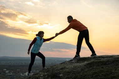 Erkek ve kadın yürüyüşçüler gün batımında dağlarda taşa tırmanmak için birbirlerine yardım ederler. Akşam vakti çift yüksek kayalara tırmanıyor. Turizm, seyahat ve sağlıklı yaşam tarzı konsepti.