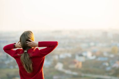 Kırmızı ceketli genç bir kadın ellerini başının arkasına koyup akşam manzarasının keyfini çıkarıyor. Rahatlama, özgürlük ve sağIık kavramı.