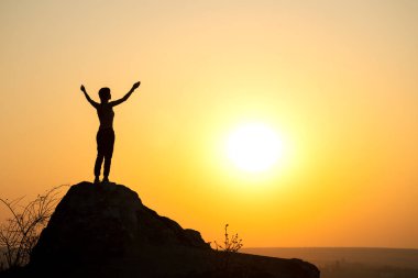 Gün batımında dağlarda büyük bir taşın üzerinde tek başına duran bir kadın yürüyüşçünün silueti. Kadın turist, akşam vakti elini yüksek kayalara kaldırıyor. Turizm, seyahat ve sağlıklı yaşam tarzı konsepti.