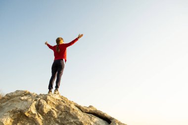 Genç kadın yürüyüşçü dağlarda büyük bir taşın üzerinde tek başına duruyor. Kadın turist sabah vakti ellerini yüksek kayalıklara kaldırıyor. Turizm, seyahat ve sağlıklı yaşam tarzı konsepti.