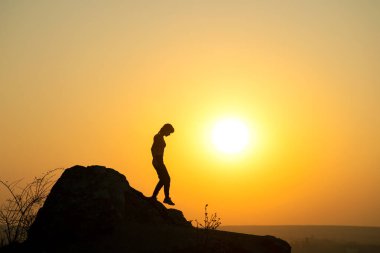 Gün batımında dağlarda büyük bir taştan inen bir kadın yürüyüşçünün silueti. Akşam vakti yüksek kayalıklarda gezen kadın turist. Turizm, seyahat ve sağlıklı yaşam tarzı konsepti.