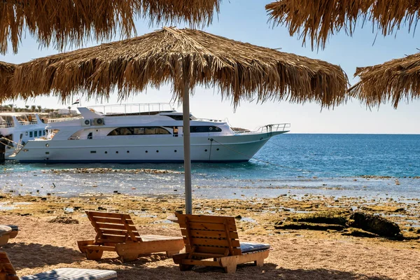 Wooden deck chairs under rough straw sun umbrella on sea beach and big white yacht ship in water near shore on sunny summer day.