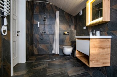 Modern tarz banyo, siyah fayanslı duvarlar, perde duş yeri ve leğen ve büyük ışıklı aynalı ahşap mobilyalar..