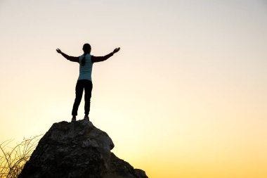 Gün batımında dağlarda büyük bir taşın üzerinde tek başına duran bir kadın yürüyüşçünün silueti. Kadın turist, akşam vakti elini yüksek kayalara kaldırıyor. Turizm, seyahat ve sağlıklı yaşam tarzı konsepti.