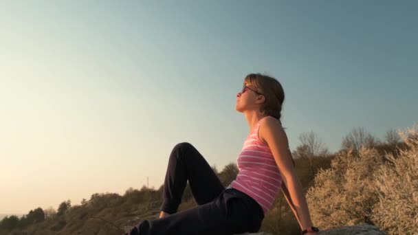 若いリラックスした女性は大きな石の上に暖かい夏の日を楽しんで屋外に座っている 女の子は瞑想し 日没時に自然の上でリラックス — ストック動画