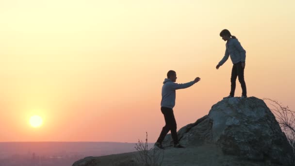男女徒步旅行者在日落时互相帮助爬上石头 黄昏的时候 夫妻二人在高高的岩石上攀爬 旅游业 旅游和健康生活方式概念 — 图库视频影像