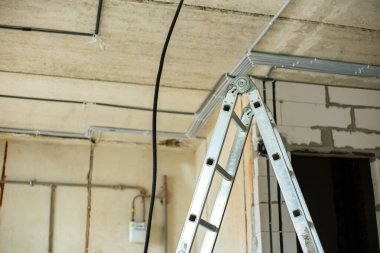 Alüminyum merdiven ve elektrik kabloları inşaat halindeki bir odanın tavanına ve duvarına yerleştirilmiş koruyucu korkuluklara yerleştirilmiş..