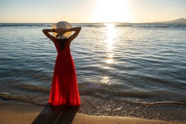 Uzun kırmızı elbise ve hasır şapka giyen genç bir kadın sahildeki deniz suyunda duruyor yaz sabahının erken saatlerinde doğan güneşin tadını çıkarıyor..