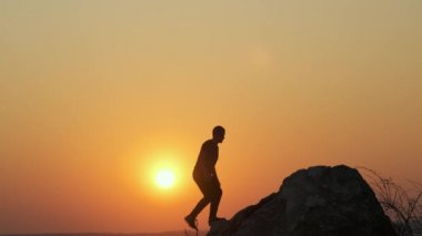 Gün batımında dağlarda tek başına büyük taşlara tırmanan bir yürüyüşçünün silueti. Erkek turist, akşam vakti elini yüksek kayalara kaldırıyor. Turizm, seyahat ve sağlıklı yaşam tarzı konsepti.