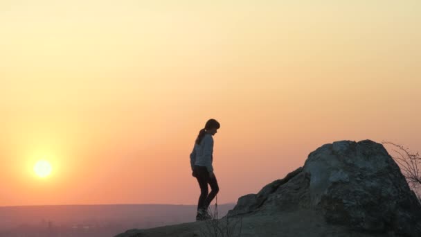 一个女徒步旅行者在日落时分独自爬上大石头的轮廓 傍晚时分 女游客举起手 在高高的岩石上滑行 旅游业 旅游和健康生活方式概念 — 图库视频影像