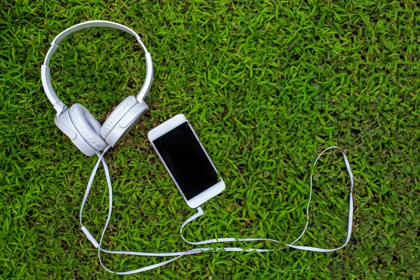 Auriculares blancos y smartphone blanco sobre hierba verde. Césped de verano con gadget personal . —  Fotos de Stock