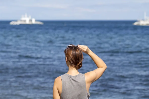 Женщина ищет морскую и белую лодку. Рыжая загорелая девушка со спины, пялящаяся на морской пейзаж — стоковое фото