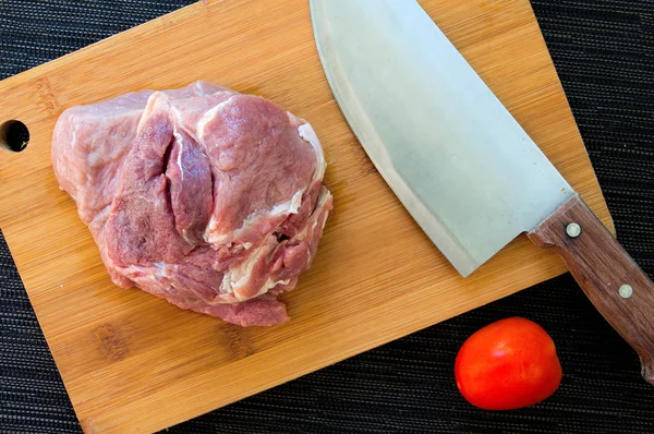 Meat and tomato on wooden board. Cutting meat by chef knife. Beef or pork for cooking. Kitchen table top view with meat and vegetables. Stainless steel knife with wood handle. Rustic kitchenware