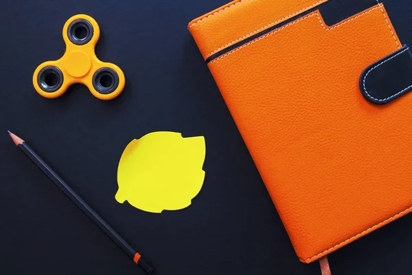 Orange notebook and spinner flat lay photo on black background. Spinner and sticker note. Autumn season flat lay with draw tools. Hipster table with blank sticker. Yellow leaf sticker with text place