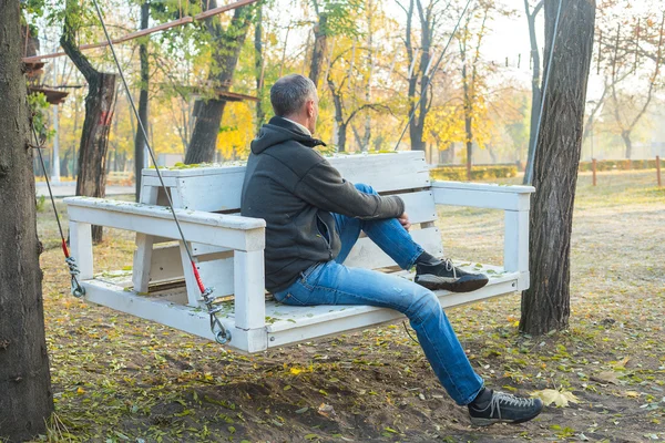 L'uomo è seduto sull'altalena nel parco autunnale Immagine Stock