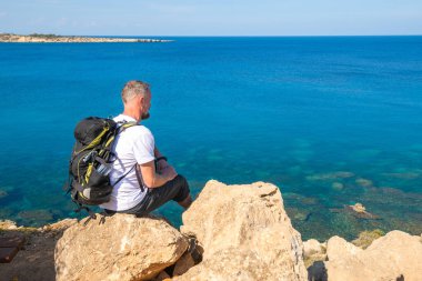 Sakallı adam sırt çantası ile kayalık kıyıda oturur 