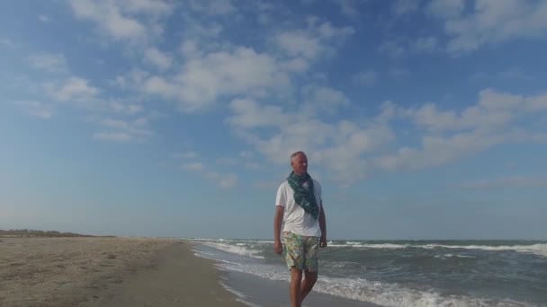Hombre sonriente vistiendo bufanda está caminando a lo largo de la línea de surf — Vídeo de stock