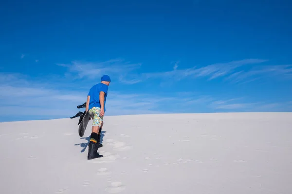 Człowiek w niebieską nasadkę ze snowboardem wspina się wydmy — Zdjęcie stockowe