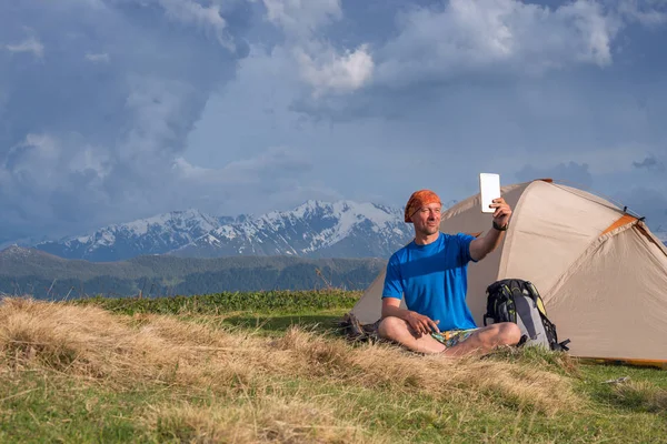 Счастливый путешественник сидит рядом с палаткой и делает селфи — стоковое фото