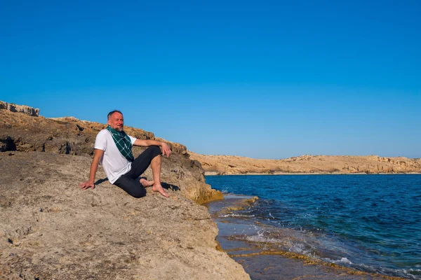 海沿いの岩のビーチに座っている髭がある旅行者 — ストック写真