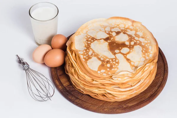 Thin pancakes on a round wooden stand with a whisk and ingredien — Stock Photo, Image