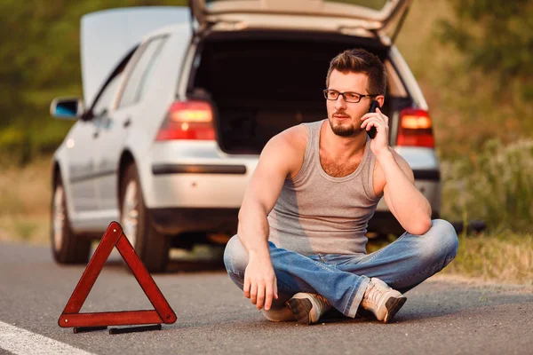 一个年轻人坐在柏油上 靠近他的破车 打电话给技术支持 道路上的损坏 — 图库照片