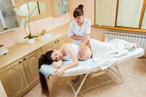 Beautiful young woman doctor massage therapist in a cosmetology room doing a massage to a pregnant girl with long hair