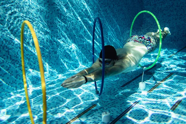 Male trainer in a cap and swimming goggles doing exercises under the water with rings