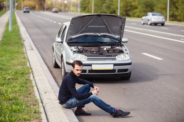 一个年轻人坐在他那辆银色汽车前面的路上 想着怎样修理它 — 图库照片