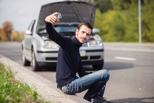 一个年轻人坐在他那辆银色汽车前面的路上 想着怎样修理它 — 图库照片