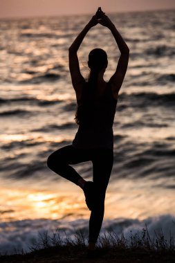 Gün batımında deniz kenarında yoga yapan bir kızın silueti.